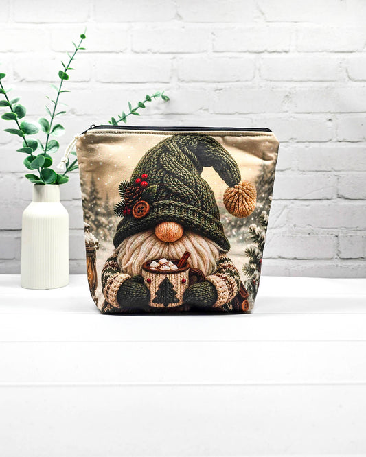 Projekttasche Isvind Größe S mit Winterwichtel, großer grüner Strickmütze, weißem Bart und einer Tasse heißem Kakao vor verschneiten Tannenbäumen. Mit Reißverschluss für kleine Strickprojekte. Handgenäht in Bayern von Nadelmikado.