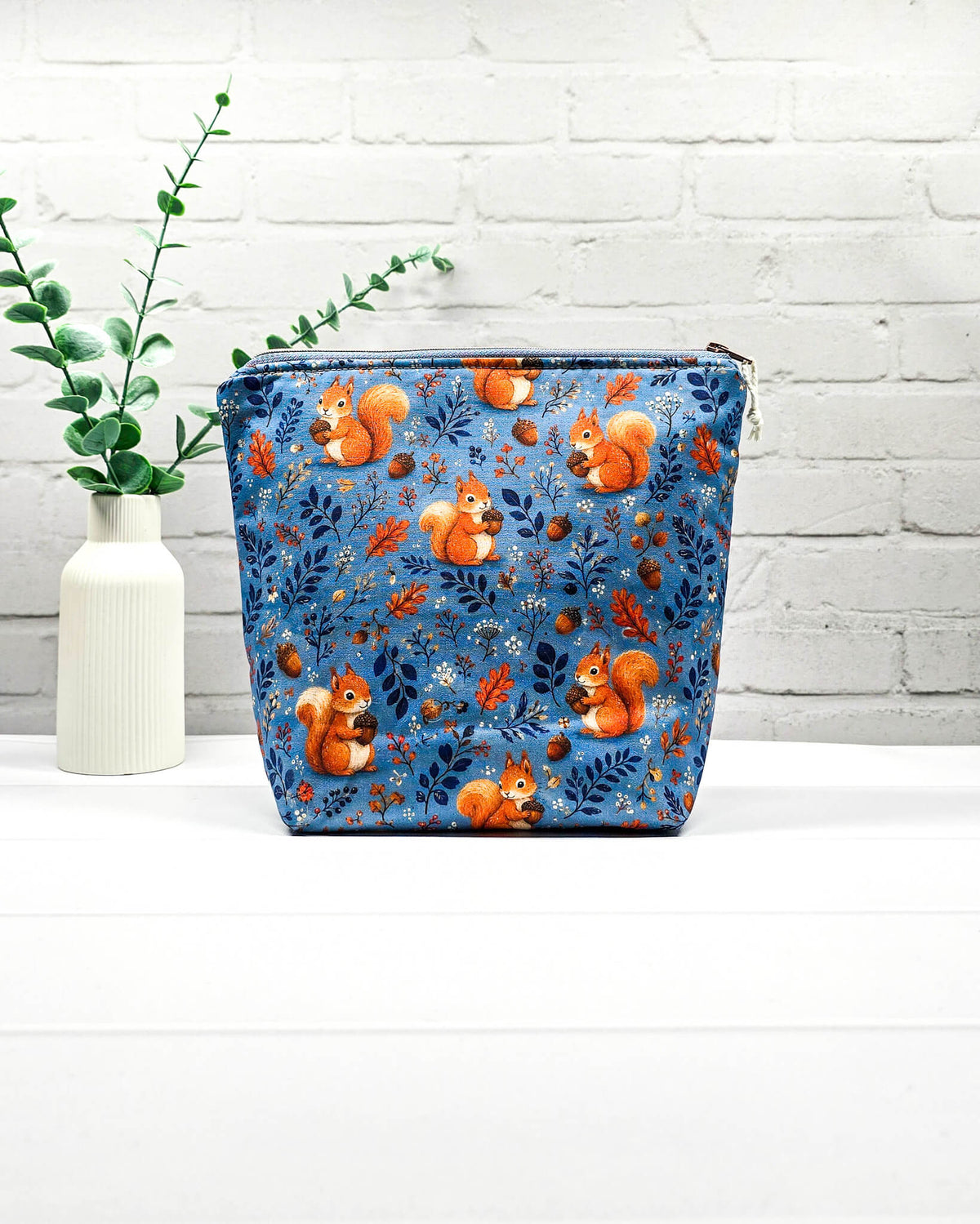 Projekttasche Größe S mit Reißverschluss aus blauem Baumwollstoff mit orangefarbenen Eichhörnchen, Eicheln und Waldzweigen zur Aufbewahrung kleiner Strickprojekte. Handgenäht in Bayern von Nadelmikado.