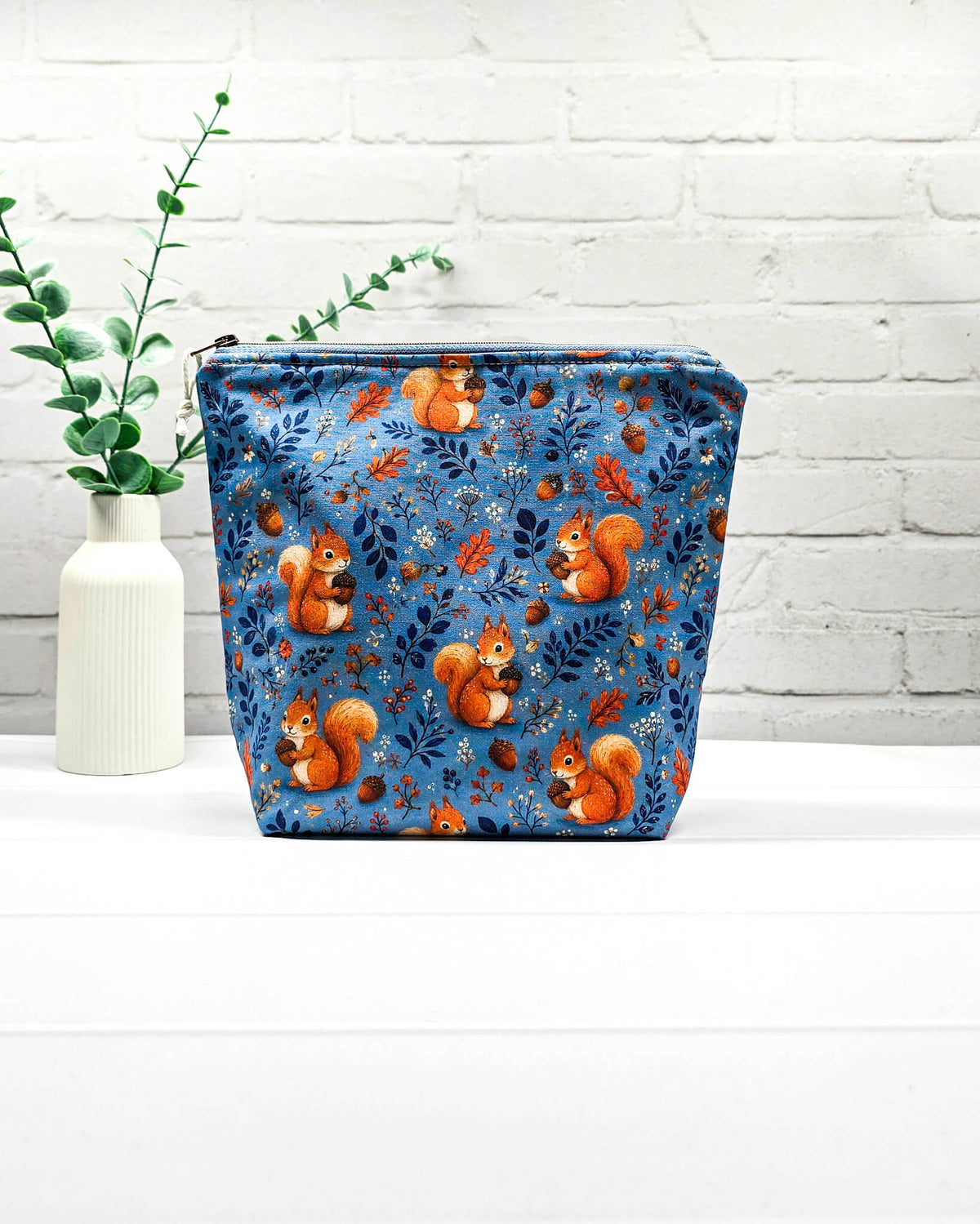 Projekttasche Größe S mit Reißverschluss aus blauem Baumwollstoff mit orangefarbenen Eichhörnchen, Eicheln und Waldzweigen zur Aufbewahrung kleiner Strickprojekte. Handgenäht in Bayern von Nadelmikado.