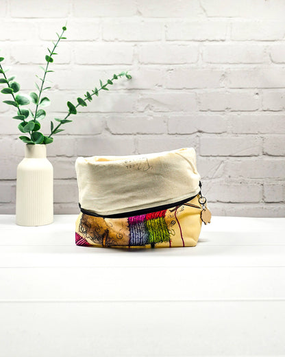 Projekttasche Isvind Größe S mit lustigem, selbst strickendem Schaf, bunter Strickmütze und Stricknadeln auf cremefarbenem Hintergrund. Mit Reißverschluss für kleine Strickprojekte. Handgenäht in Bayern von Nadelmikado.