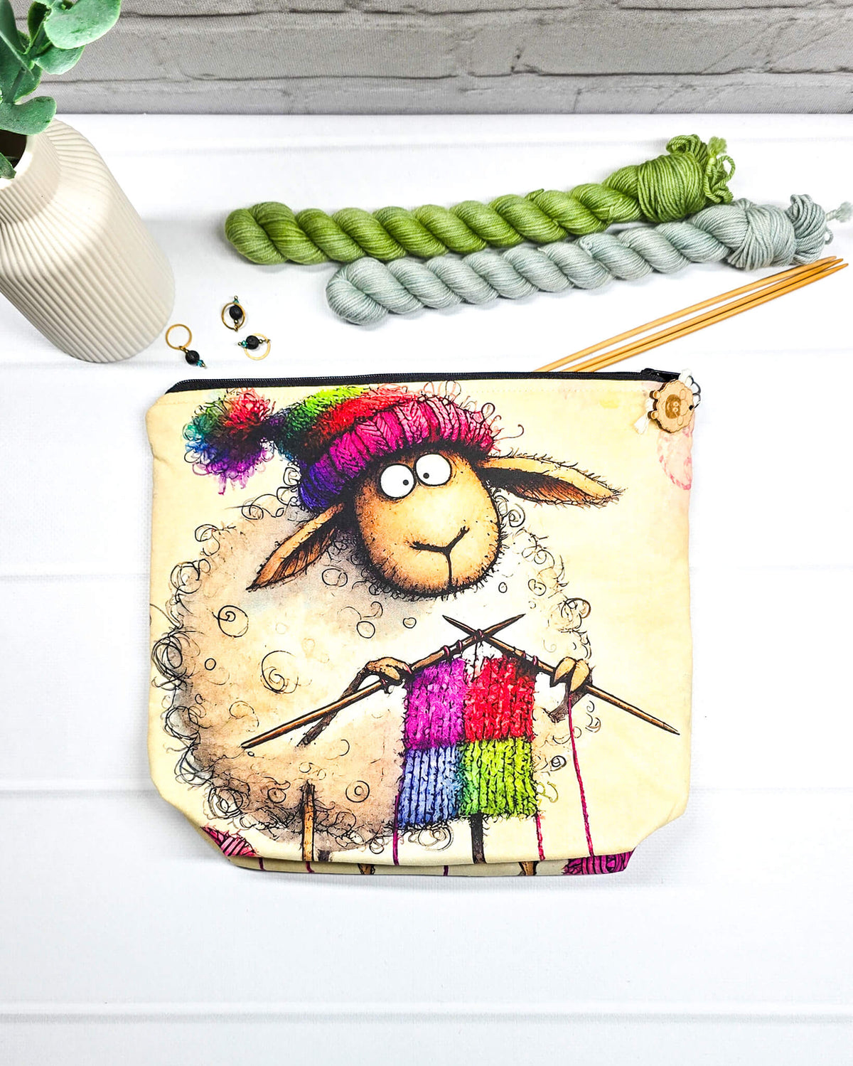 Projekttasche Isvind Größe S mit lustigem, selbst strickendem Schaf, bunter Strickmütze und Stricknadeln auf cremefarbenem Hintergrund. Mit Reißverschluss für kleine Strickprojekte. Handgenäht in Bayern von Nadelmikado.