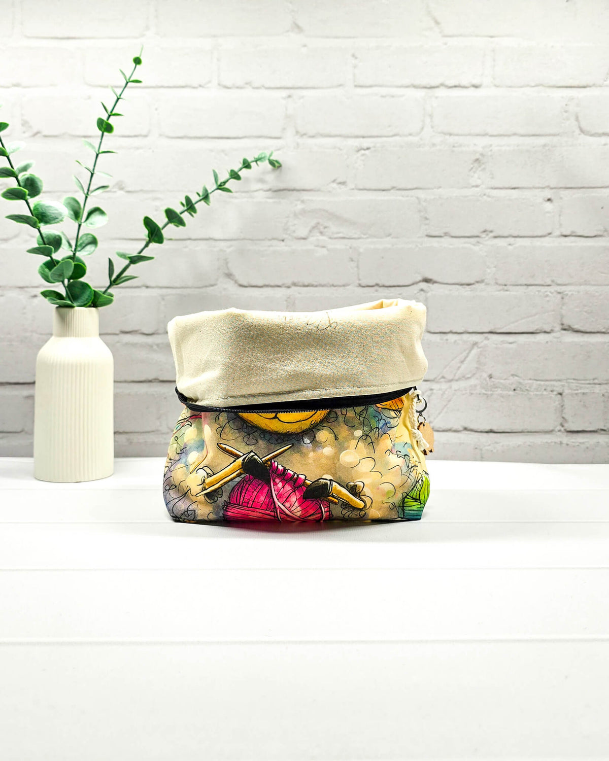 Projekttasche Isvind Größe S mit lustigem Schafmotiv, buntem Lockenkopf, Stricknadeln und pinker Wolle auf hellem Hintergrund. Mit Reißverschluss für kleine Strickprojekte. Handgenäht in Bayern von Nadelmikado.
