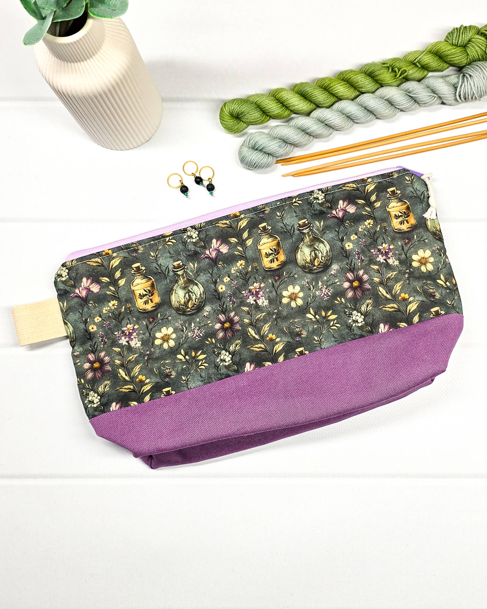 Projekttasche Isvind S mit Elixierfläschchen und Wildblumen auf dunkelgrünem Grund, lilafarbenem Canvas-Boden und lila Reißverschluss für Sockenwolle. Handgenäht in Bayern von Nadelmikado.