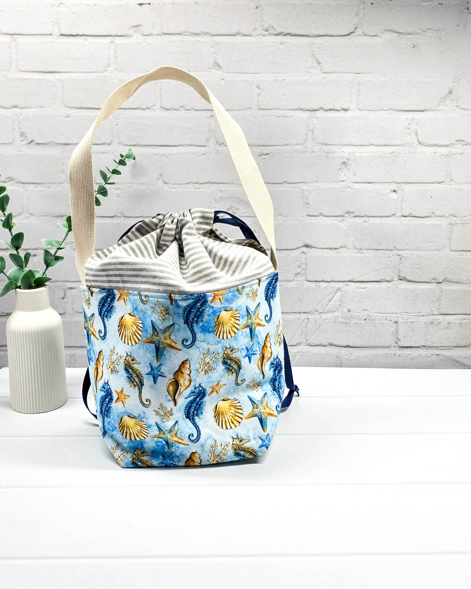 Projekttasche Juna S mit Seepferdchen- und Muschel-Motiv auf Blau, grau-weiß gestreiftem Kordelzugbereich und einem langen, hellen Schultergurt. Handgenäht in Bayern von Nadelmikado. 