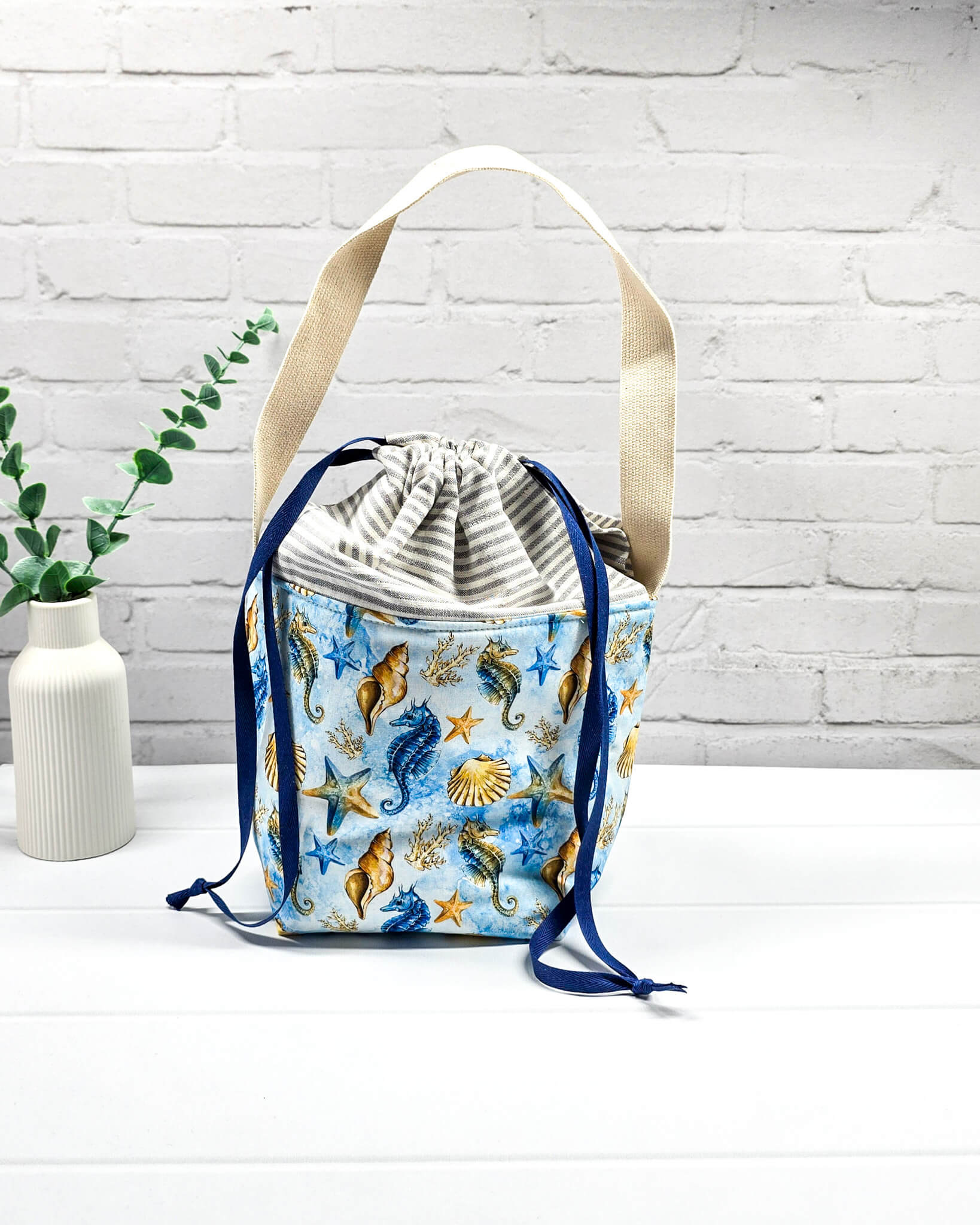 Projekttasche Juna S mit Seepferdchen- und Muschel-Motiv auf Blau, grau-weiß gestreiftem Kordelzugbereich und einem langen, hellen Schultergurt. Handgenäht in Bayern von Nadelmikado. 
