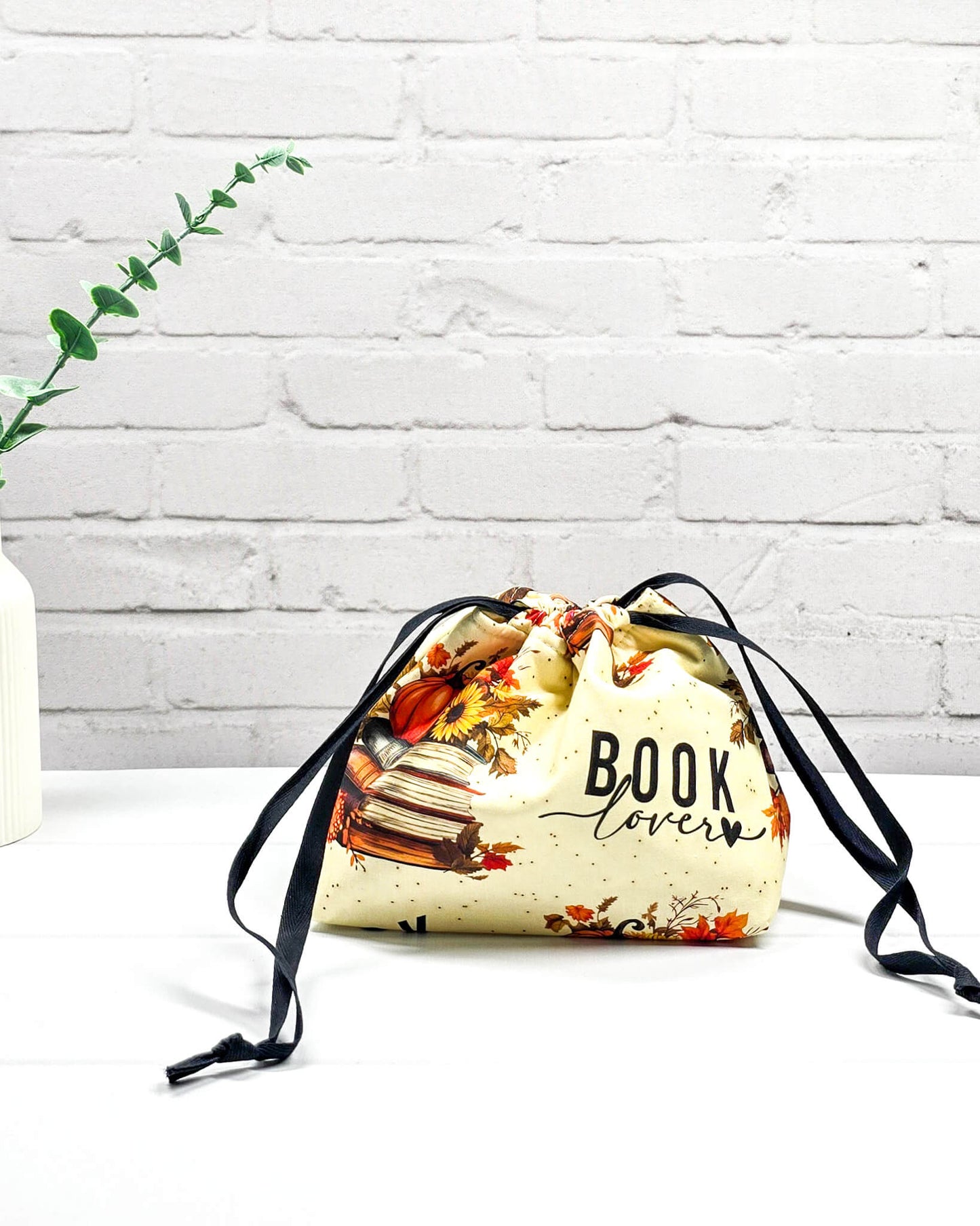 Projekttasche in Größe S für Sockenprojekte bis 100 g Wolle. Der Kinchaku-Verschluss bewahrt dein Strickzeug sicher auf. Motiv mit "Book Lover"-Schriftzug, Buchstapeln und herbstlichen Blumen. Handgenäht in Bayern von Nadelmikado