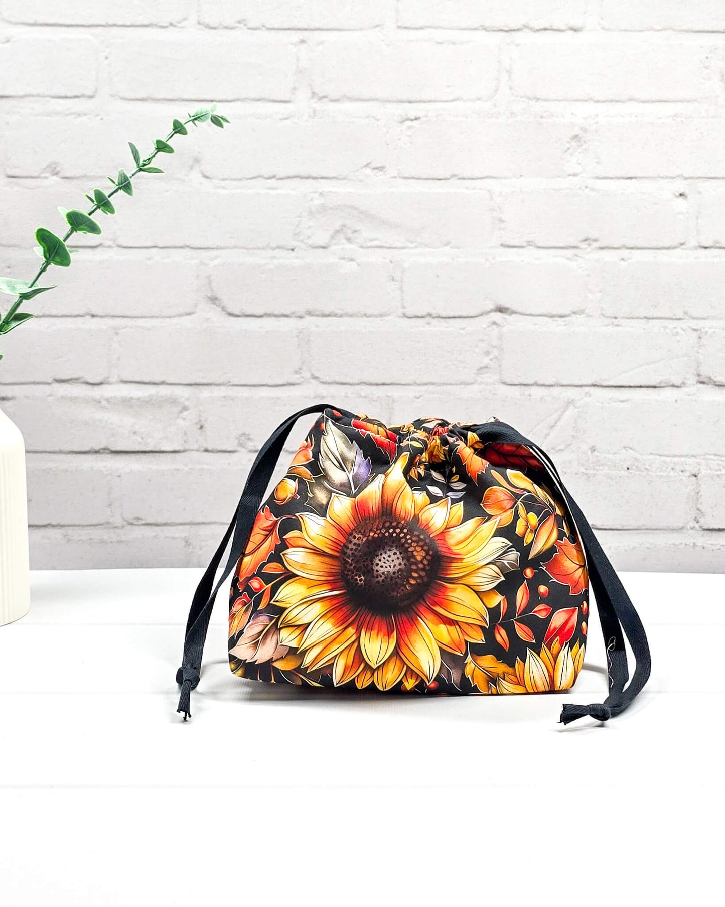 Projekttasche in Größe S für Sockenprojekte bis 100 g Wolle. Kinchaku-Verschluss sichert das Strickzeug. Motiv mit großer Sonnenblume und Herbstlaub auf dunklem Stoff. Handgenäht in Bayern von Nadelmikado. 