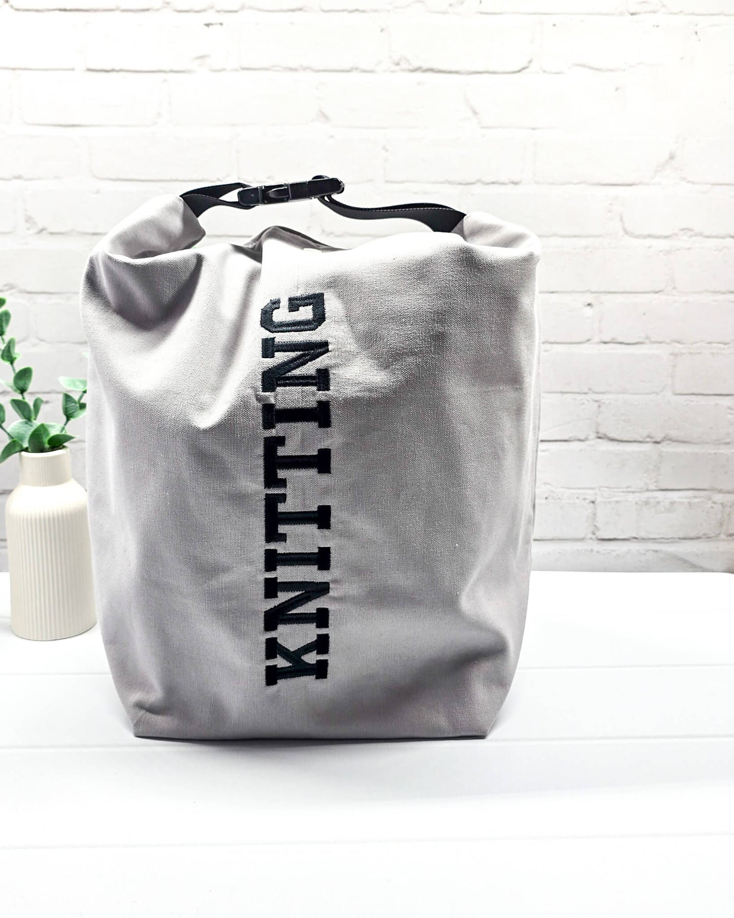 Projekttasche Rika im Roll-Top-Design aus hellgrauem Canvas mit vertikalem, schwarzem Schriftzug „KNITTING“ und schwarzem Gurtband-Verschluss. Handgenäht in Bayern von Nadelmikado