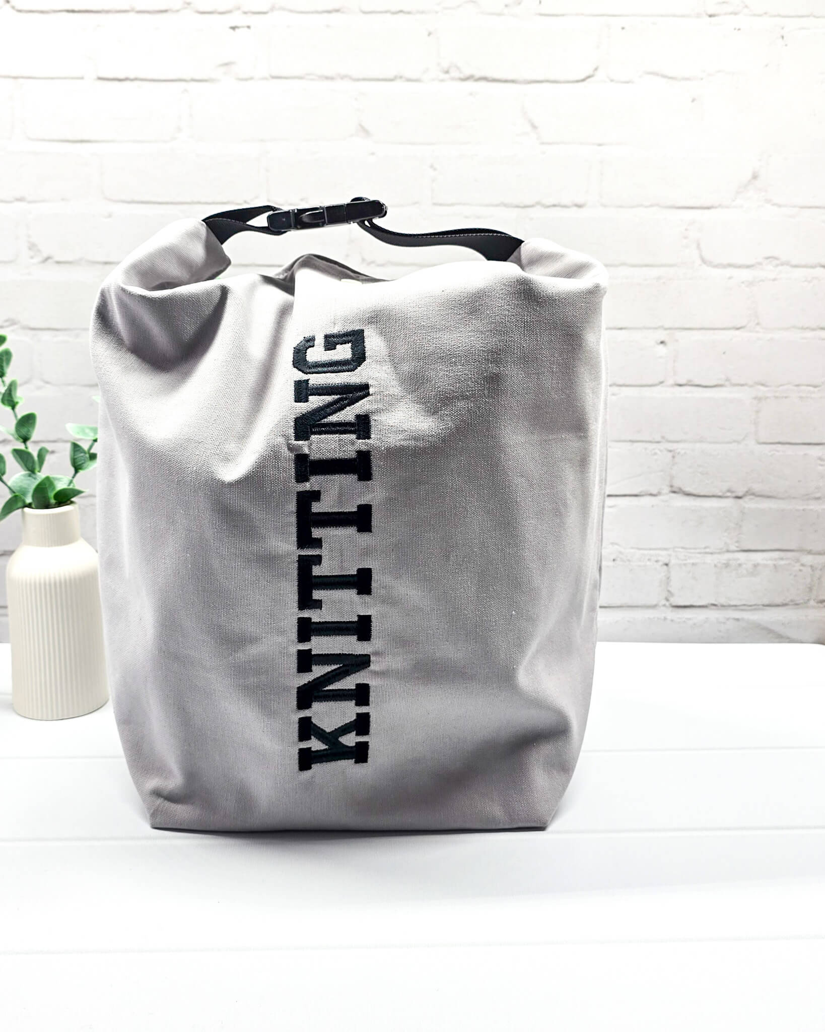 Projekttasche Rika im Roll-Top-Design aus hellgrauem Canvas mit vertikalem, schwarzem Schriftzug „KNITTING“ und schwarzem Gurtband-Verschluss. Handgenäht in Bayern von Nadelmikado