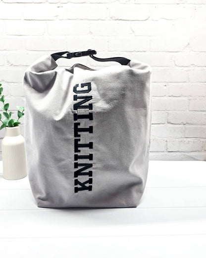 Projekttasche Rika im Roll-Top-Design aus hellgrauem Canvas mit vertikalem, schwarzem Schriftzug „KNITTING“ und schwarzem Gurtband-Verschluss. Handgenäht in Bayern von Nadelmikado