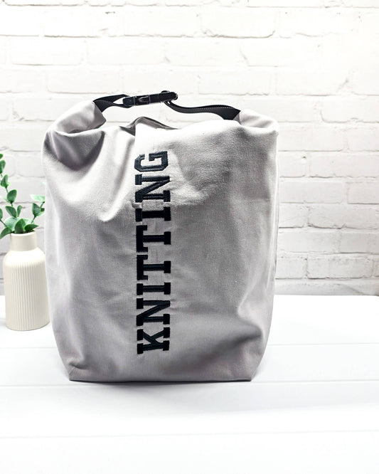 Projekttasche Rika im Roll-Top-Design aus hellgrauem Canvas mit vertikalem, schwarzem Schriftzug „KNITTING“ und schwarzem Gurtband-Verschluss. Handgenäht in Bayern von Nadelmikado