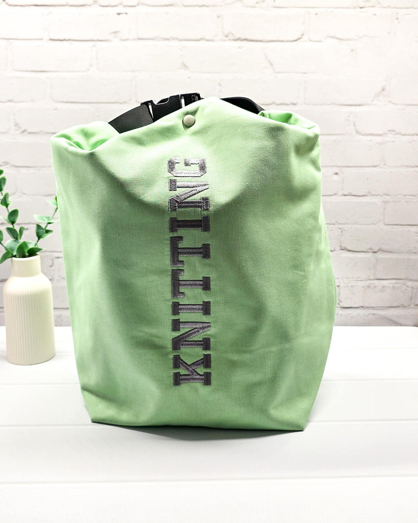 Projekttasche Rika im Roll-Top-Design aus hellgrünem Canvas mit vertikalem, dunkelgrauem Schriftzug „KNITTING“ und schwarzem Gurtband-Verschluss. Handgenäht in Bayern von Nadelmikado. 