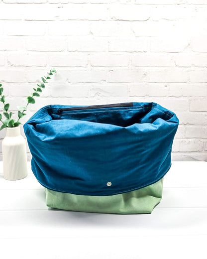 Projekttasche Rika im Roll-Top-Design aus hellgrünem Canvas mit vertikalem, dunkelgrünem Schriftzug „KNITTING“ und petrolfarbenem Gurtband-Verschluss. Handgenäht in Bayern von Nadelmikado. 