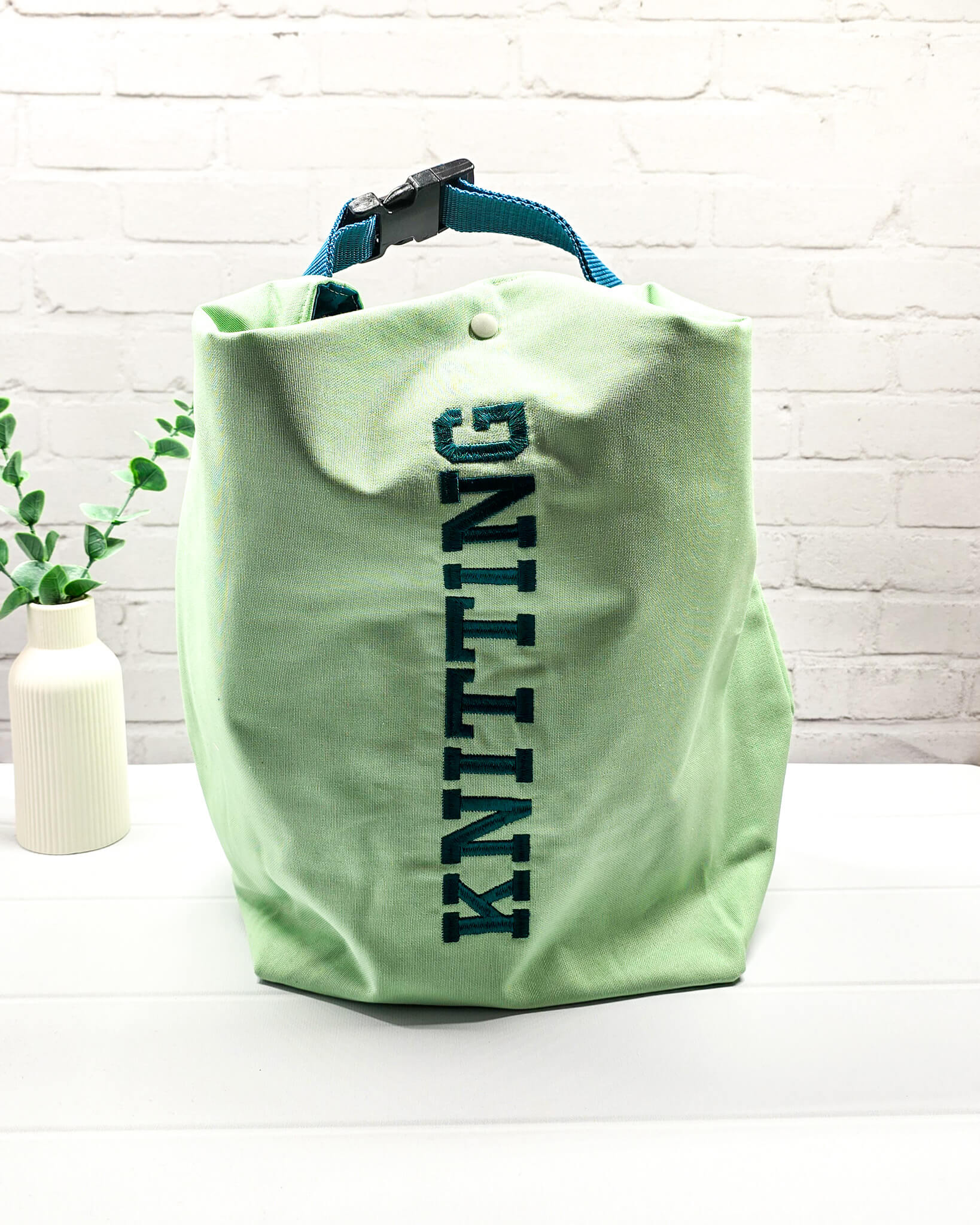 Projekttasche Rika im Roll-Top-Design aus hellgrünem Canvas mit vertikalem, dunkelgrünem Schriftzug „KNITTING“ und petrolfarbenem Gurtband-Verschluss. Handgenäht in Bayern von Nadelmikado. 