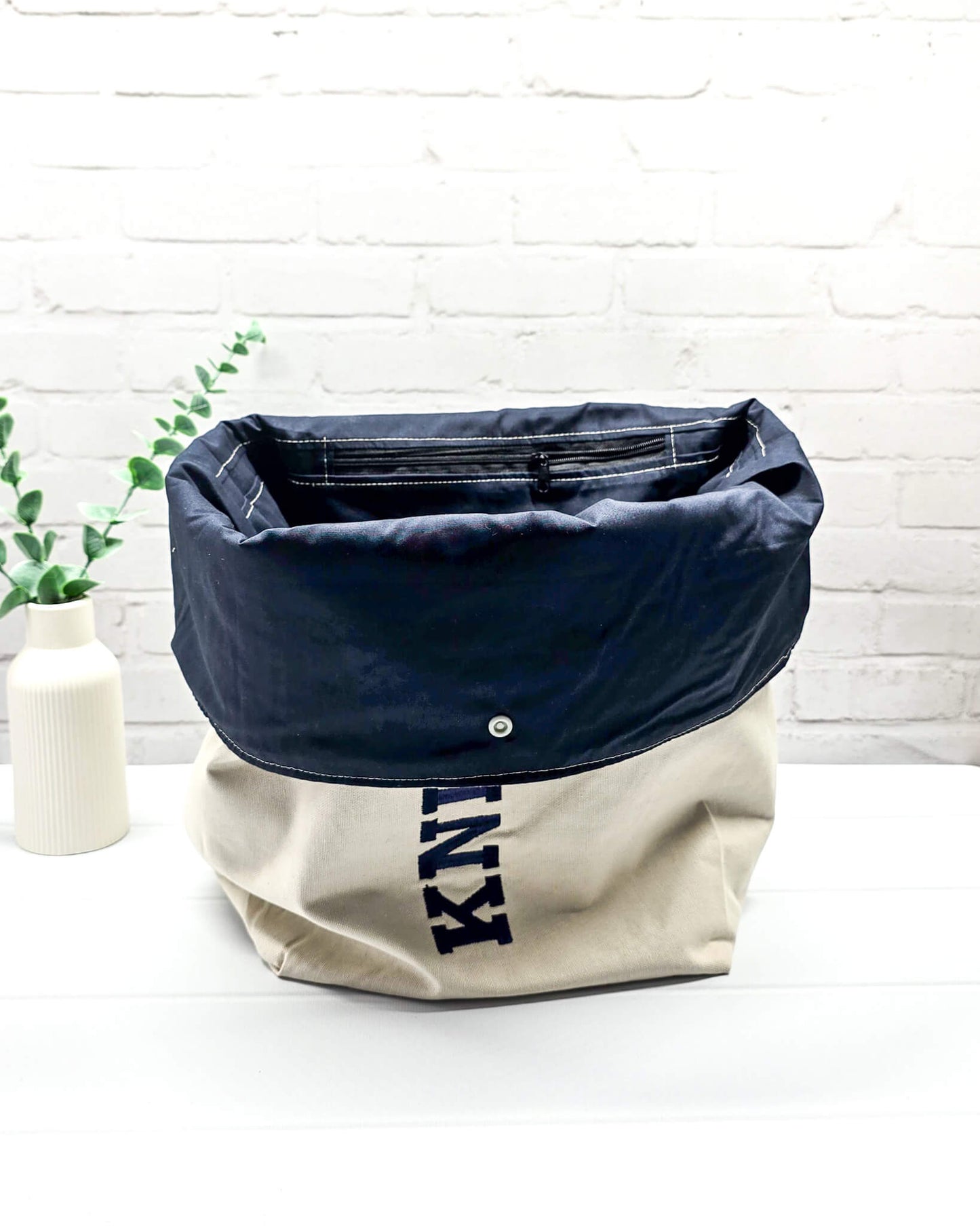 Projekttasche Rika im Roll-Top-Design aus naturfarbenem Canvas mit vertikalem, dunkelblauem Schriftzug „KNITTING“ und blauem Gurtband-Verschluss. Handgenäht in Bayern von Nadelmikado. 