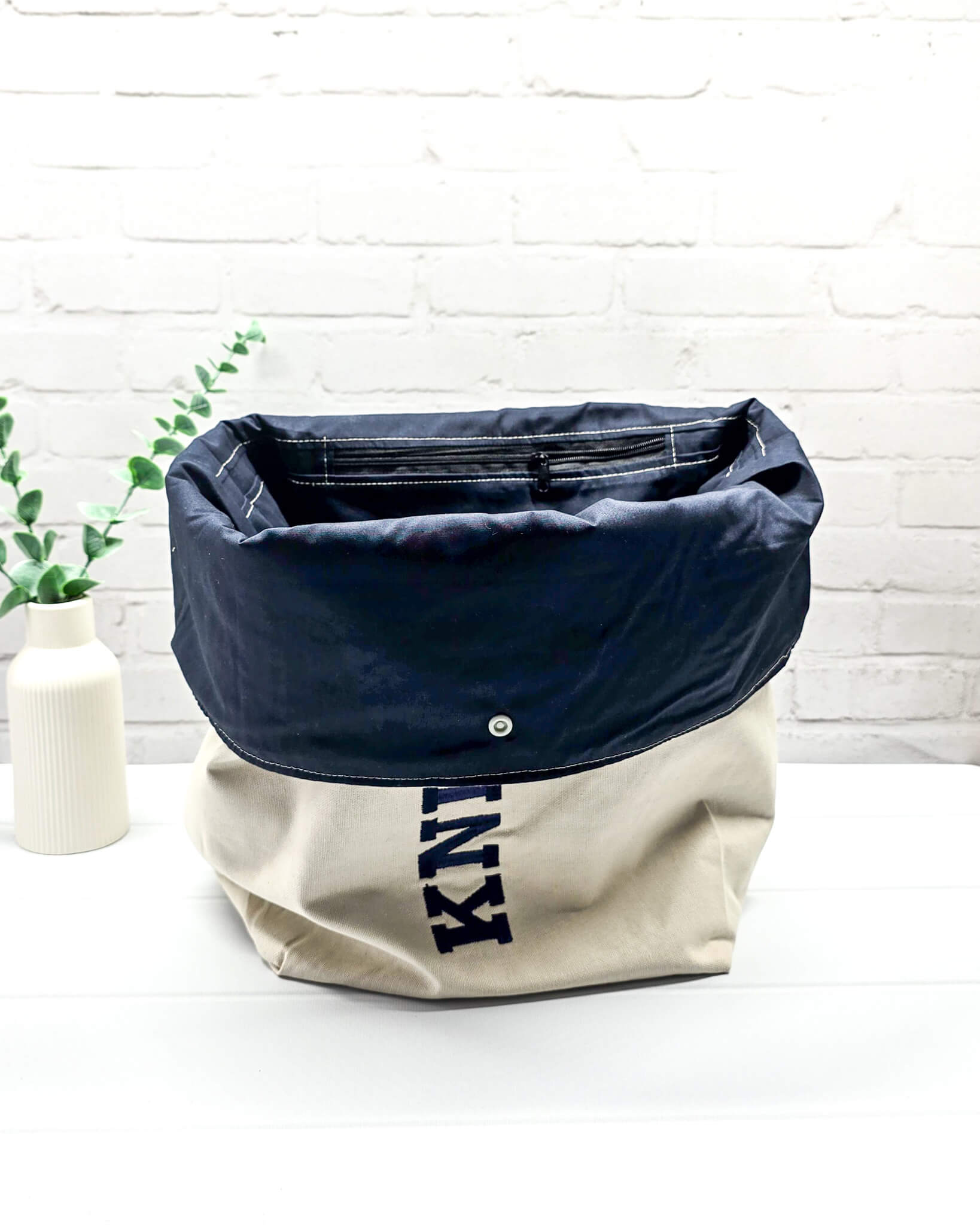 Projekttasche Rika im Roll-Top-Design aus naturfarbenem Canvas mit vertikalem, dunkelblauem Schriftzug „KNITTING“ und blauem Gurtband-Verschluss. Handgenäht in Bayern von Nadelmikado. 