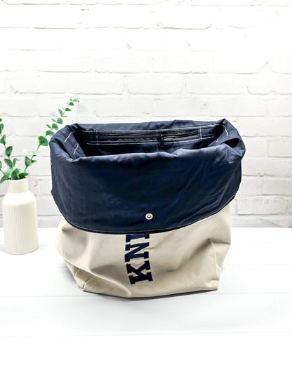 Projekttasche Rika im Roll-Top-Design aus naturfarbenem Canvas mit vertikalem, dunkelblauem Schriftzug „KNITTING“ und blauem Gurtband-Verschluss. Handgenäht in Bayern von Nadelmikado. 