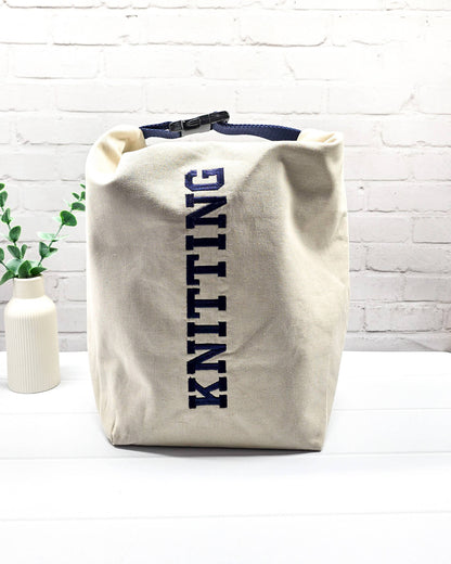 Projekttasche Rika im Roll-Top-Design aus naturfarbenem Canvas mit vertikalem, dunkelblauem Schriftzug „KNITTING“ und blauem Gurtband-Verschluss. Handgenäht in Bayern von Nadelmikado. 
