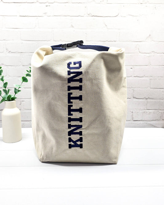 Projekttasche Rika im Roll-Top-Design aus naturfarbenem Canvas mit vertikalem, dunkelblauem Schriftzug „KNITTING“ und blauem Gurtband-Verschluss. Handgenäht in Bayern von Nadelmikado. 