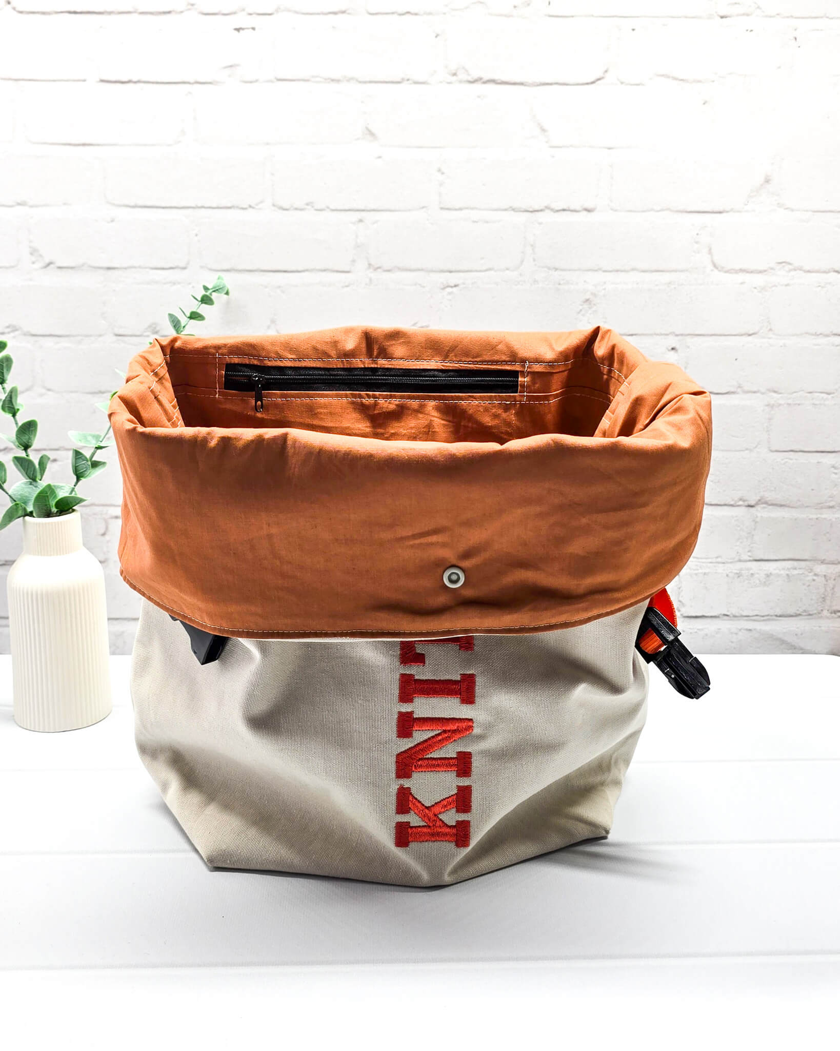 Projekttasche Rika im Roll-Top-Design aus naturfarbenem Canvas mit vertikalem, orangerotem Schriftzug „KNITTING“ und orangem Gurtband-Verschluss. Handgenäht in Bayern von Nadelmikado. 