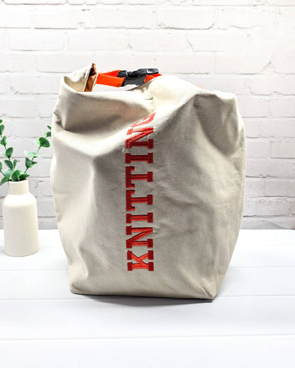Projekttasche Rika im Roll-Top-Design aus naturfarbenem Canvas mit vertikalem, orangerotem Schriftzug „KNITTING“ und orangem Gurtband-Verschluss. Handgenäht in Bayern von Nadelmikado. 