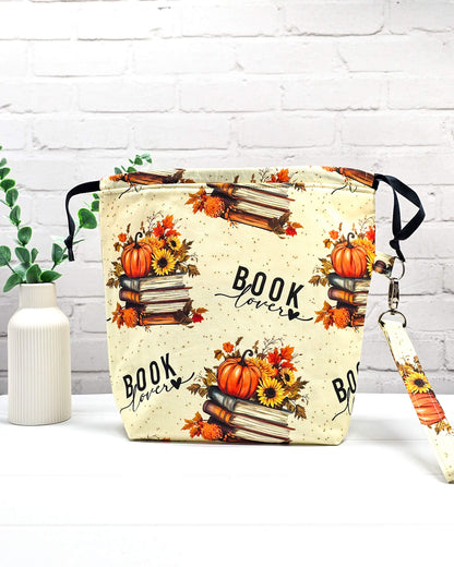 Projekttasche Tuva M aus cremefarbenem Baumwollstoff mit Bücherstapeln, Kürbissen, dem Schriftzug „BOOK lover“ und abnehmbarer Handschlaufe. Handgenäht in Bayern von Nadelmikado. 