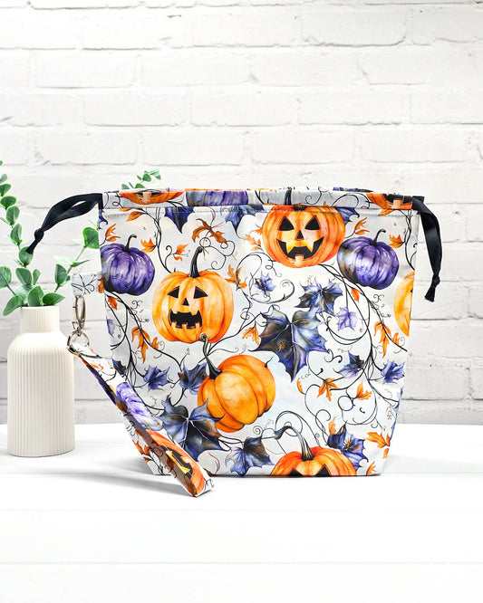 Projekttasche Tuva M aus weißem Baumwollstoff mit Halloween-Kürbissen, lilafarbenen Blättern, schwarzem Kordelzug und abnehmbarer Handschlaufe. Handgenäht in Bayern von Nadelmikado