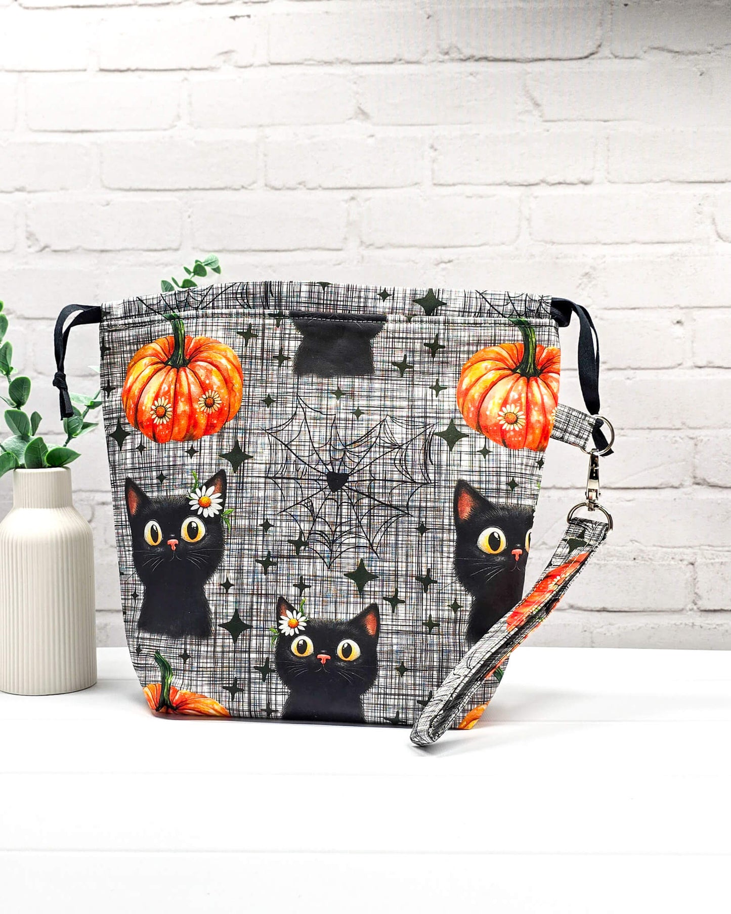 Projekttasche Tuva M aus grau-schwarz kariertem Baumwollstoff mit schwarzen Halloween-Katzen, Kürbissen und einer abnehmbaren Handschlaufe. Handgenäht in Bayern von Nadelmikado. 