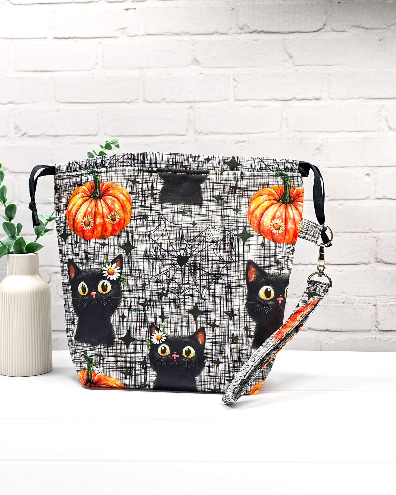 Projekttasche Tuva M aus grau-schwarz kariertem Baumwollstoff mit schwarzen Halloween-Katzen, Kürbissen und einer abnehmbaren Handschlaufe. Handgenäht in Bayern von Nadelmikado. 