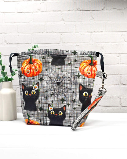 Projekttasche Tuva M aus grau-schwarz kariertem Baumwollstoff mit schwarzen Halloween-Katzen, Kürbissen und einer abnehmbaren Handschlaufe. Handgenäht in Bayern von Nadelmikado. 