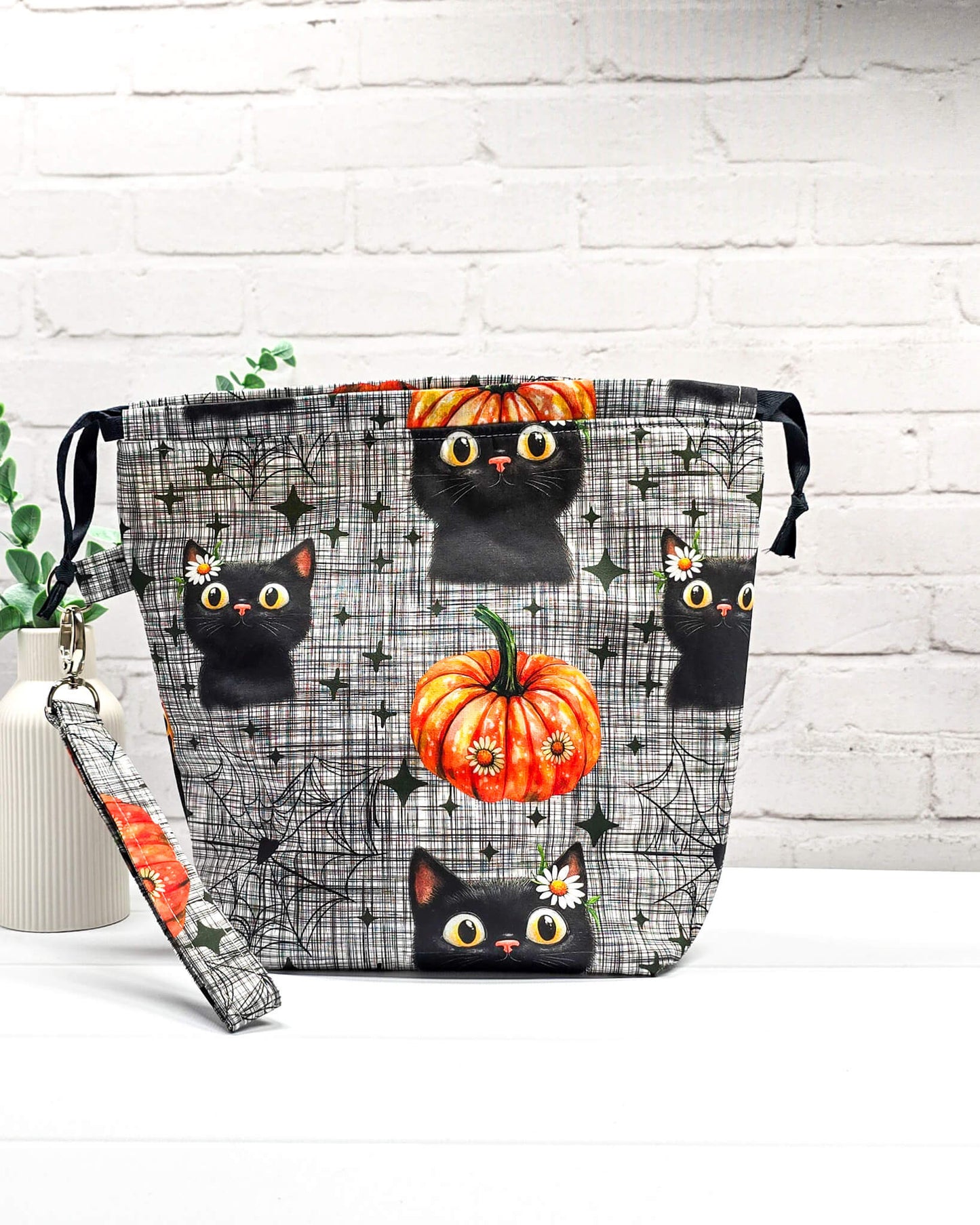 Projekttasche Tuva M aus grau-schwarz kariertem Baumwollstoff mit schwarzen Halloween-Katzen, Kürbissen und einer abnehmbaren Handschlaufe. Handgenäht in Bayern von Nadelmikado. 