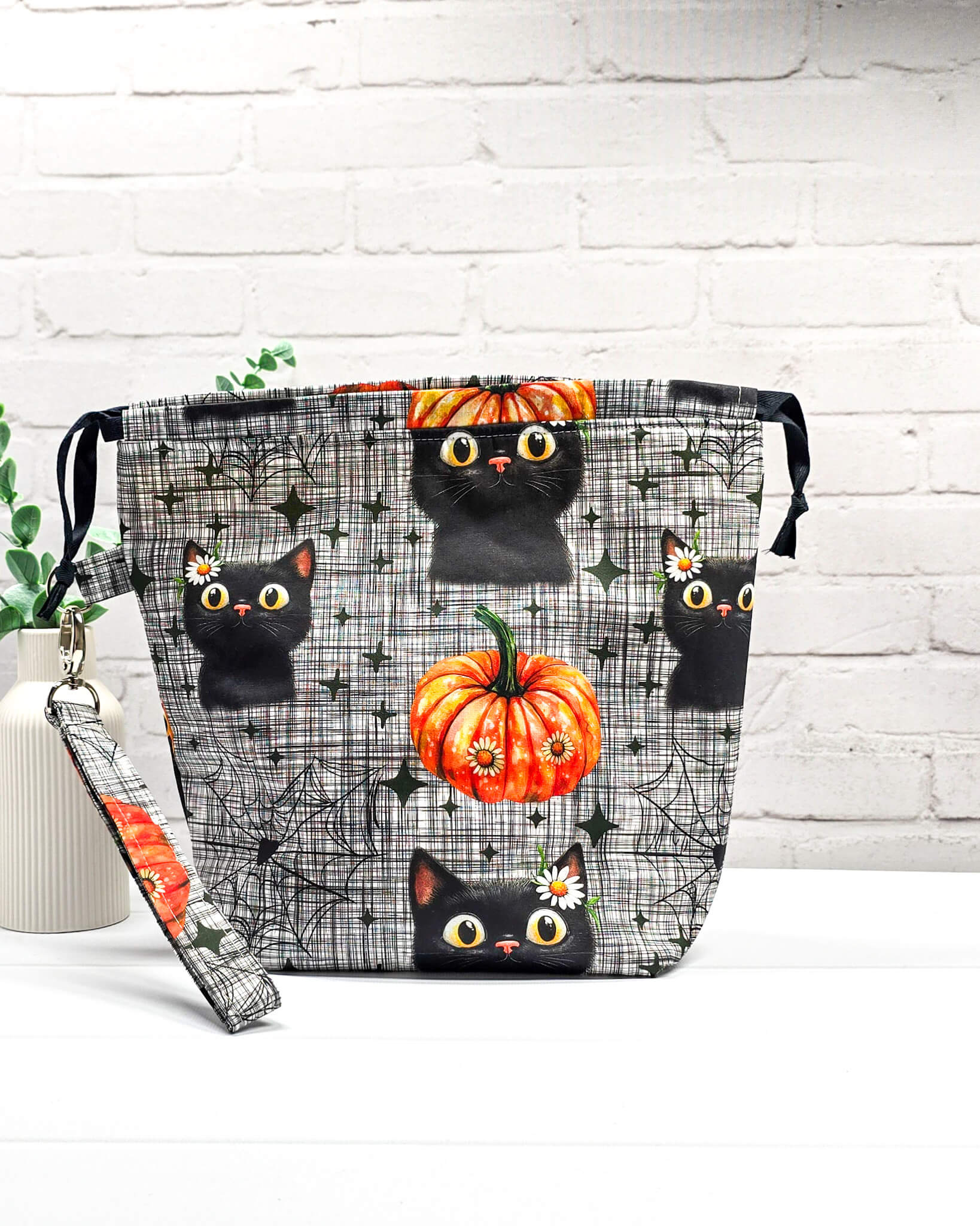 Projekttasche Tuva M aus grau-schwarz kariertem Baumwollstoff mit schwarzen Halloween-Katzen, Kürbissen und einer abnehmbaren Handschlaufe. Handgenäht in Bayern von Nadelmikado. 