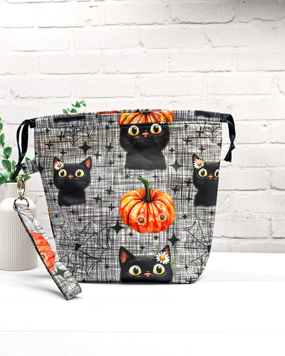 Projekttasche Tuva M aus grau-schwarz kariertem Baumwollstoff mit schwarzen Halloween-Katzen, Kürbissen und einer abnehmbaren Handschlaufe. Handgenäht in Bayern von Nadelmikado. 