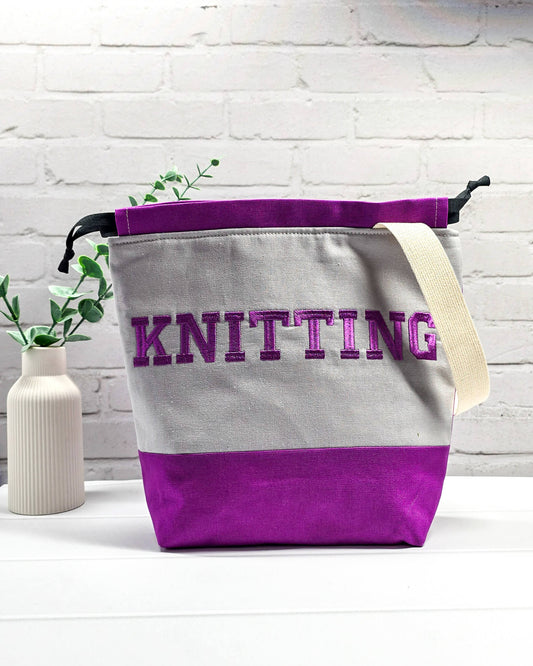 Bestickte Projekttasche Tuva M aus grauem und lila Canvas-Stoff mit violettem Schriftzug KNITTING, schwarzem Kordelzug und cremefarbenem Tragegurt. Bietet Platz für 400g Wolle und schützt das Strickzeug vor Verheddern. Handgenäht in Bayern von Nadelmikado.