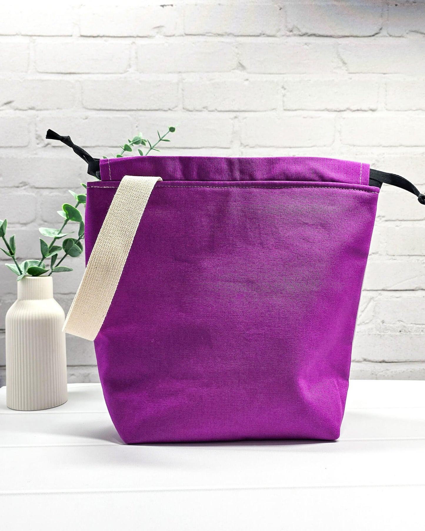 Bestickte Projekttasche Tuva M aus grauem und lila Canvas-Stoff mit violettem Schriftzug KNITTING, schwarzem Kordelzug und cremefarbenem Tragegurt. Bietet Platz für 400g Wolle und schützt das Strickzeug vor Verheddern. Handgenäht in Bayern von Nadelmikado.