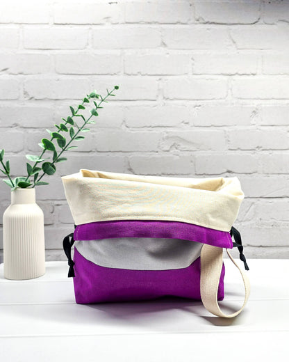 Bestickte Projekttasche Tuva M aus grauem und lila Canvas-Stoff mit violettem Schriftzug KNITTING, schwarzem Kordelzug und cremefarbenem Tragegurt. Bietet Platz für 400g Wolle und schützt das Strickzeug vor Verheddern. Handgenäht in Bayern von Nadelmikado.