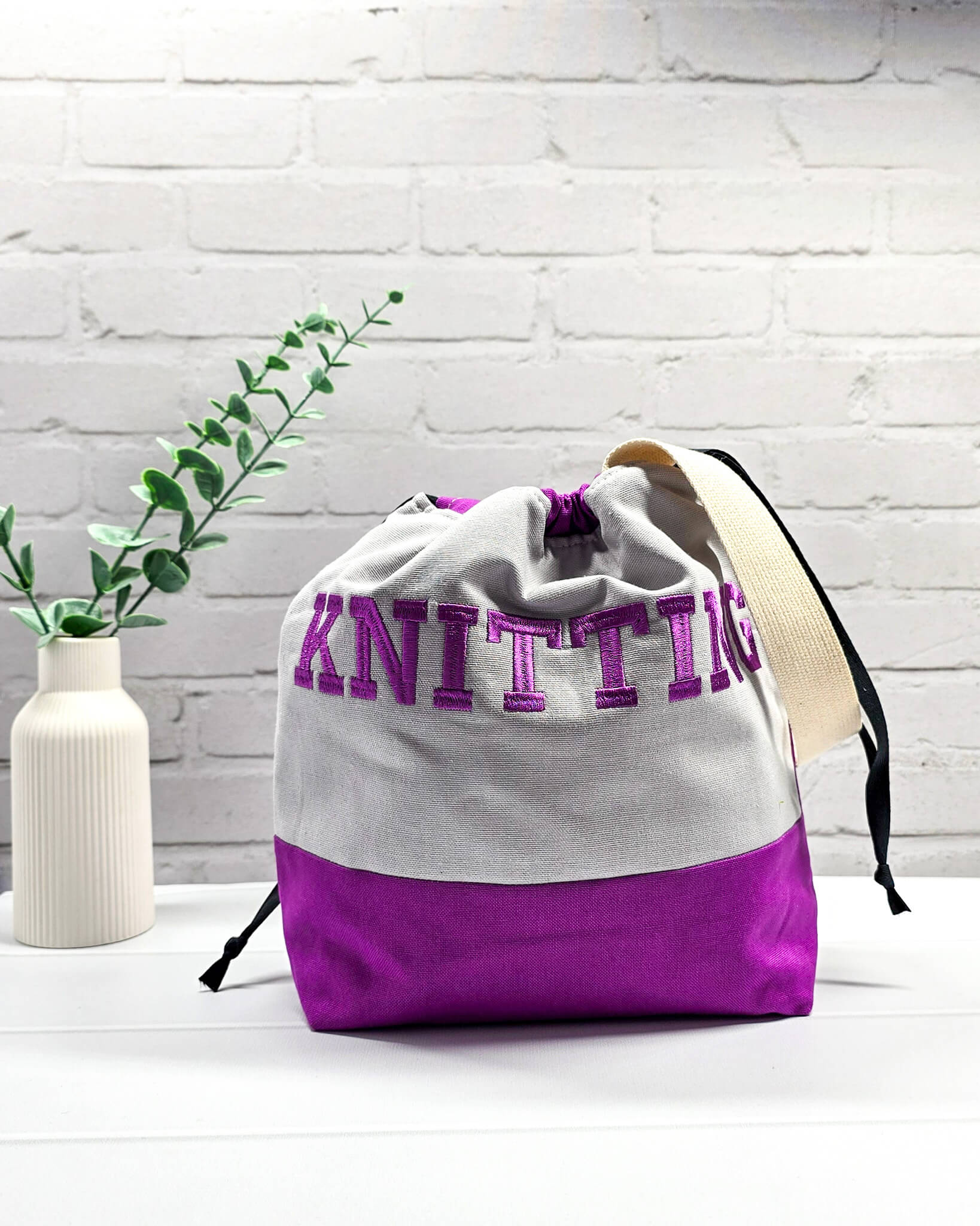 Bestickte Projekttasche Tuva M aus grauem und lila Canvas-Stoff mit violettem Schriftzug KNITTING, schwarzem Kordelzug und cremefarbenem Tragegurt. Bietet Platz für 400g Wolle und schützt das Strickzeug vor Verheddern. Handgenäht in Bayern von Nadelmikado.