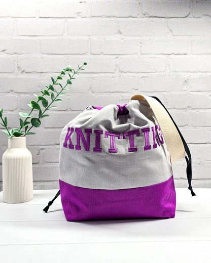 Bestickte Projekttasche Tuva M aus grauem und lila Canvas-Stoff mit violettem Schriftzug KNITTING, schwarzem Kordelzug und cremefarbenem Tragegurt. Bietet Platz für 400g Wolle und schützt das Strickzeug vor Verheddern. Handgenäht in Bayern von Nadelmikado.