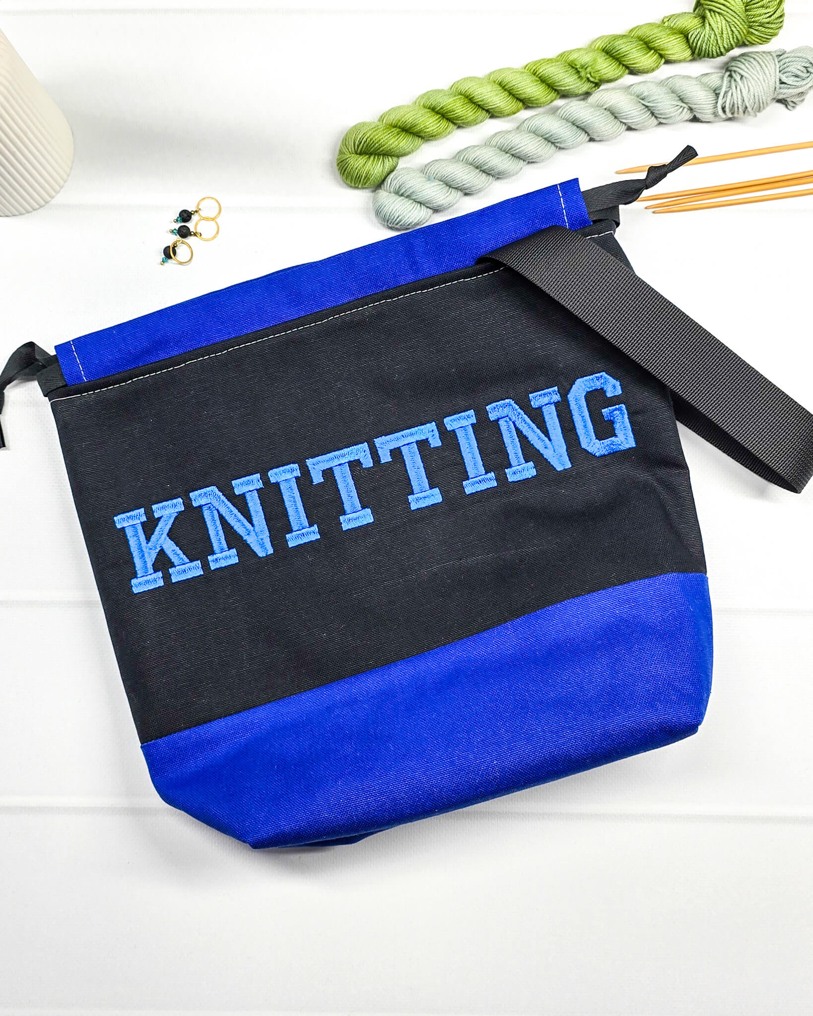 Bestickte Projekttasche Tuva M aus schwarzem und blauem Canvas-Stoff mit blauem Schriftzug KNITTING, schwarzem Kordelzug und schwarzem Tragegurt. Bietet Platz für 400g Wolle und schützt das Strickzeug vor Verheddern. Handgenäht in Bayern von Nadelmikado.