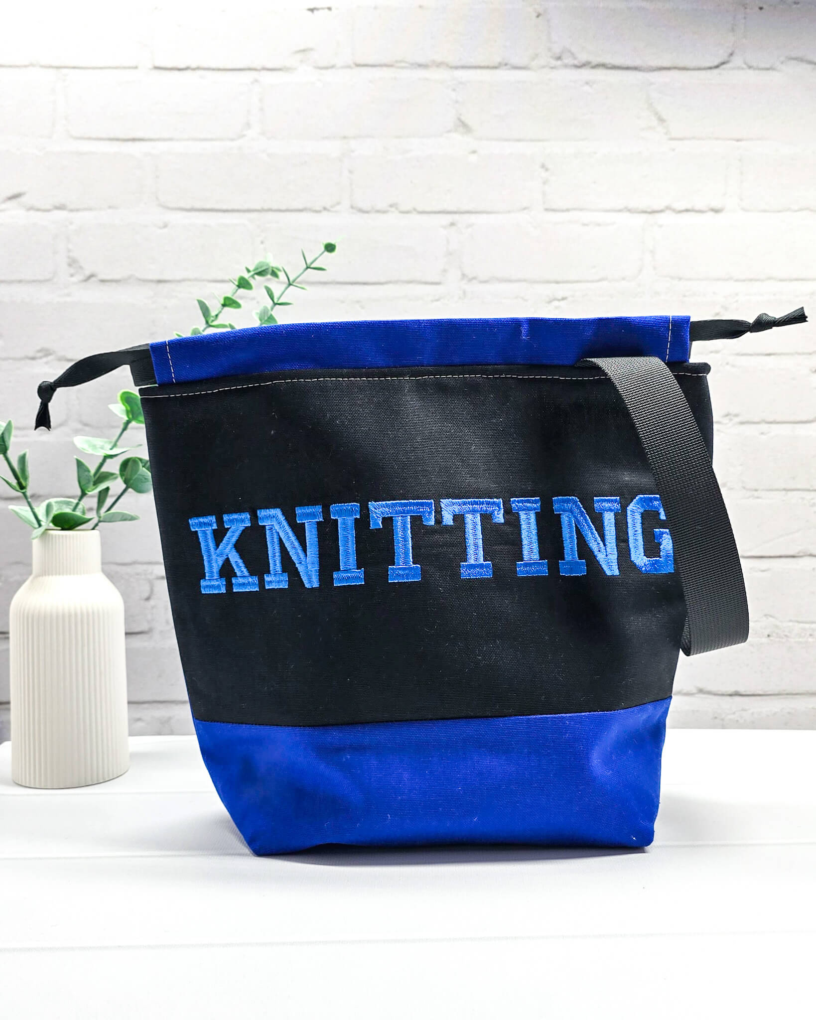 Bestickte Projekttasche Tuva M aus schwarzem und blauem Canvas-Stoff mit blauem Schriftzug KNITTING, schwarzem Kordelzug und schwarzem Tragegurt. Bietet Platz für 400g Wolle und schützt das Strickzeug vor Verheddern. Handgenäht in Bayern von Nadelmikado.