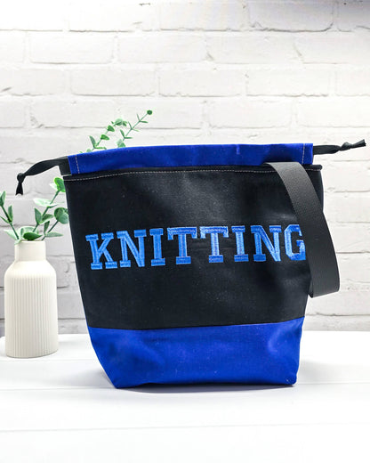 Bestickte Projekttasche Tuva M aus schwarzem und blauem Canvas-Stoff mit blauem Schriftzug KNITTING, schwarzem Kordelzug und schwarzem Tragegurt. Bietet Platz für 400g Wolle und schützt das Strickzeug vor Verheddern. Handgenäht in Bayern von Nadelmikado.