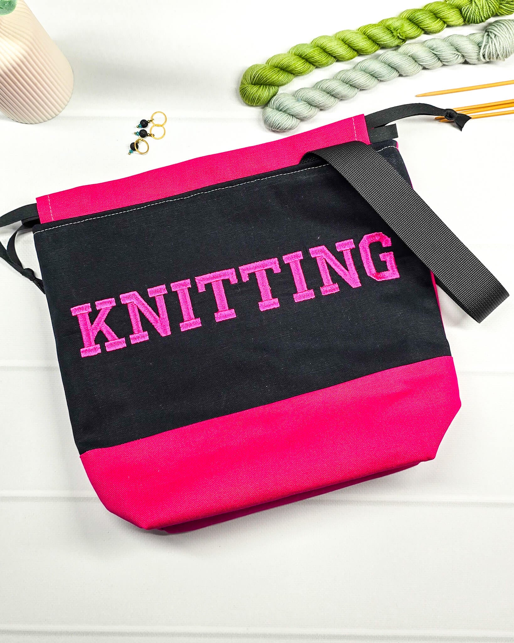 Bestickte Projekttasche Tuva M aus schwarzem und pinkem Canvas-Stoff mit pinkem Schriftzug KNITTING, schwarzem Kordelzug und schwarzem Tragegurt. Bietet Platz für 400g Wolle. Handgenäht in Bayern von Nadelmikado.