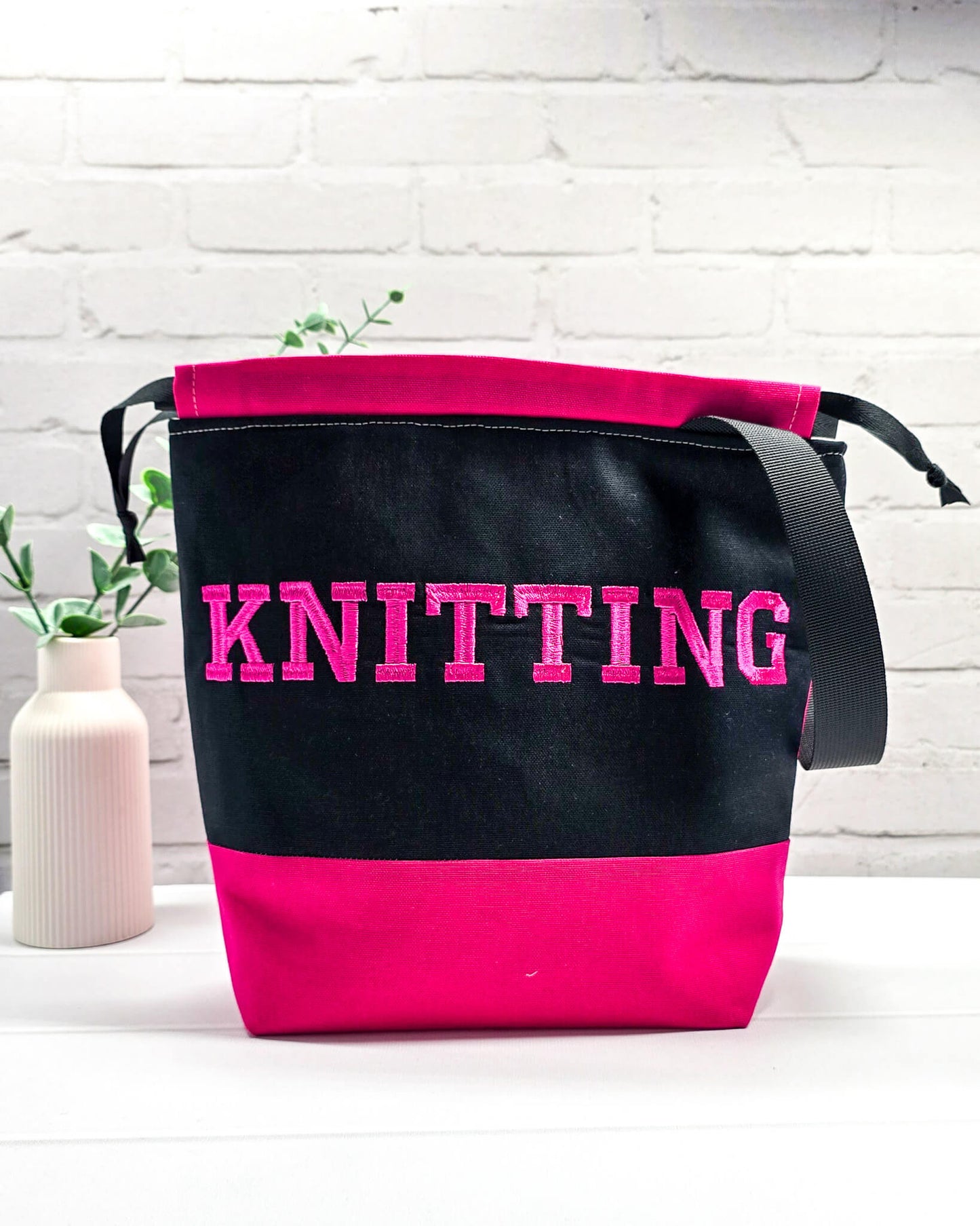 Bestickte Projekttasche Tuva M aus schwarzem und pinkem Canvas-Stoff mit pinkem Schriftzug KNITTING, schwarzem Kordelzug und schwarzem Tragegurt. Bietet Platz für 400g Wolle. Handgenäht in Bayern von Nadelmikado.