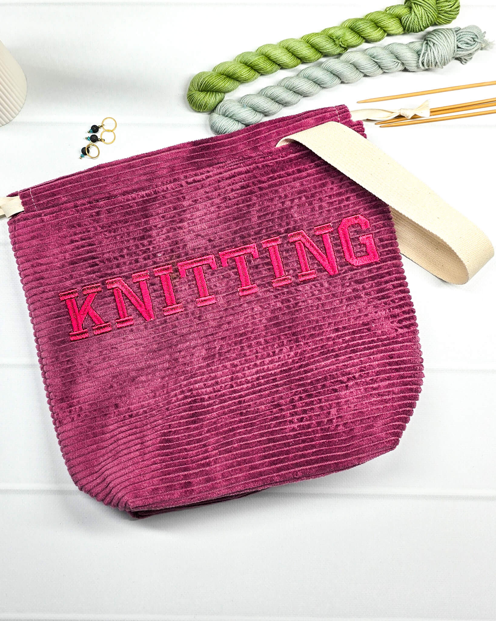 Bestickte Projekttasche Tuva M aus beerefarbenem Breitcord mit dem Schriftzug KNITTING, Kordelzug und cremefarbenem Tragegurt. Bietet Platz für 400g Wolle und schützt das Strickzeug vor Verheddern. Handgenäht in Bayern von Nadelmikado.