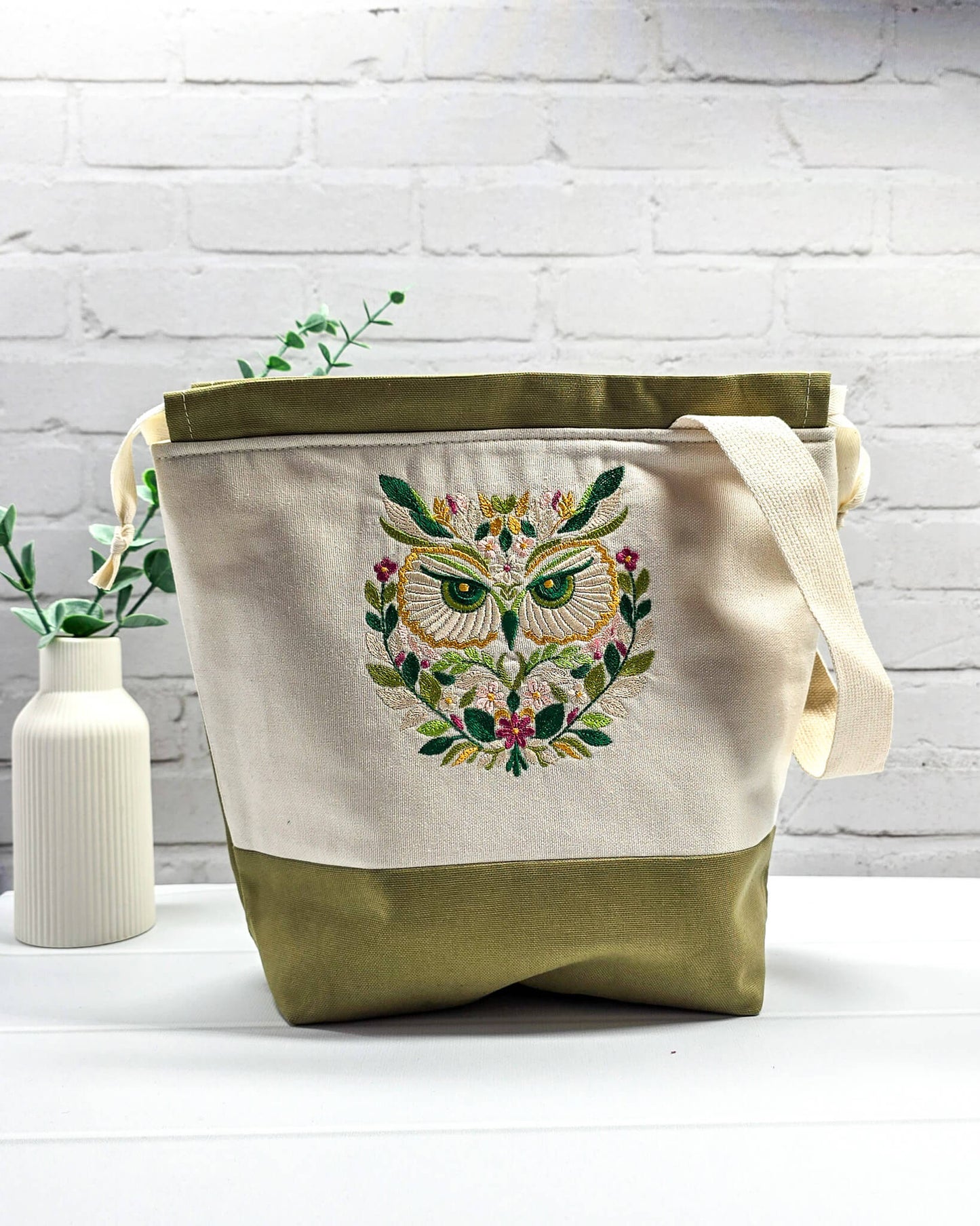 Projekttasche Tuva M aus hellem Canvas mit gesticktem Eulenmotiv, olivgrünem Boden, Kordelzug und Tragegurt. Bietet Platz für 400g Wolle. Handgenäht in Bayern von Nadelmikado.