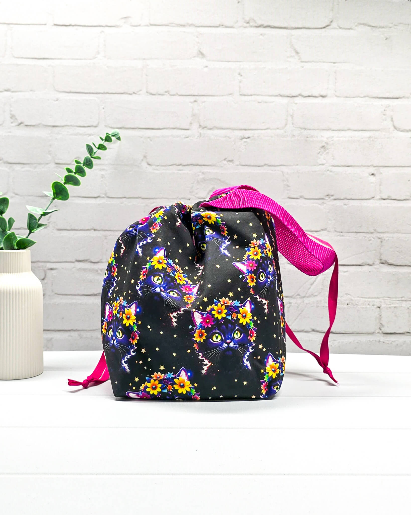 Projekttasche Tuva M aus schwarzem Baumwollstoff mit bunten Blumen-Katzen, pinkem Kordelzug und pinker Trageschlaufe. Handgenäht in Bayern von Nadelmikado. 