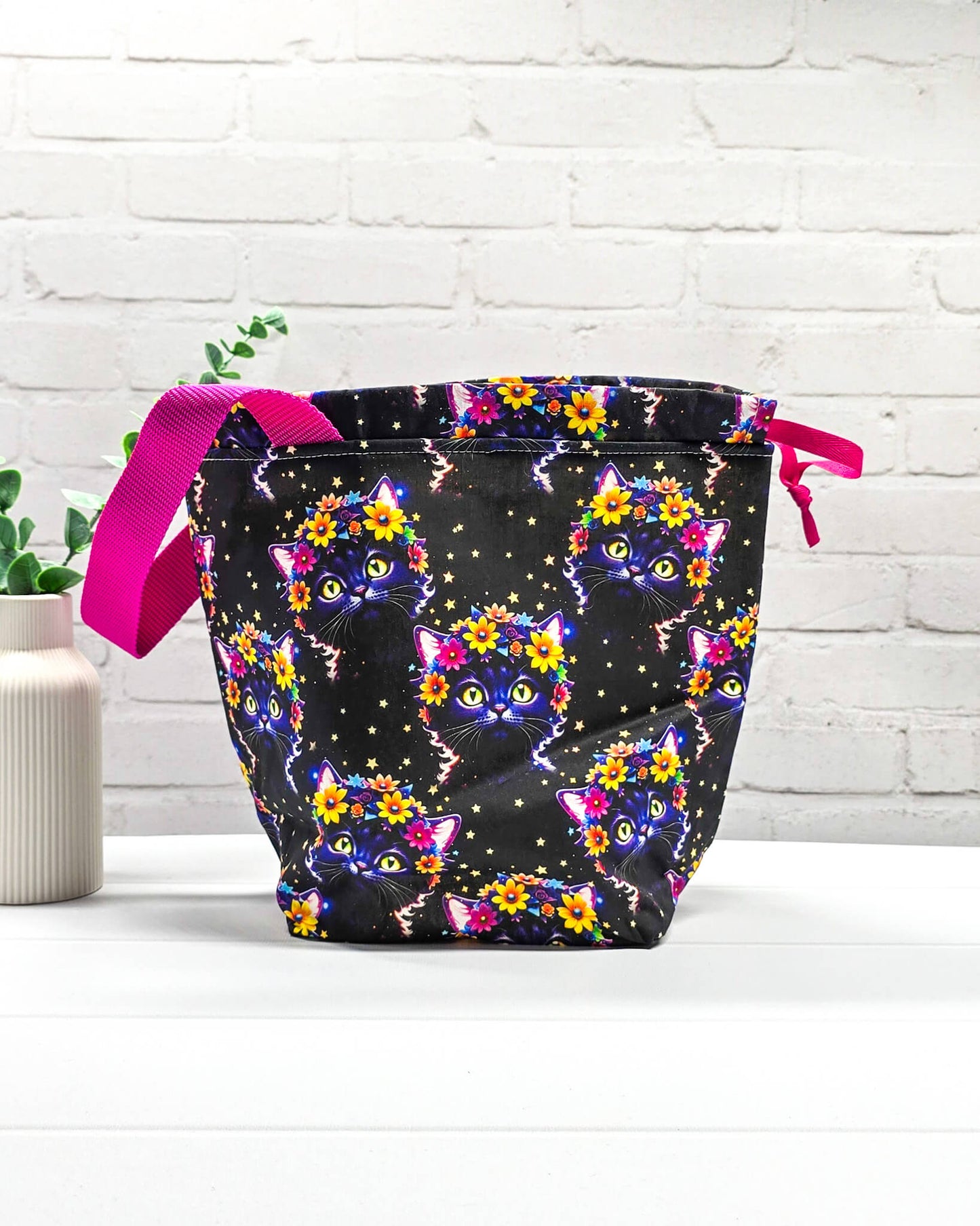 Projekttasche Tuva M aus schwarzem Baumwollstoff mit bunten Blumen-Katzen, pinkem Kordelzug und pinker Trageschlaufe. Handgenäht in Bayern von Nadelmikado. 