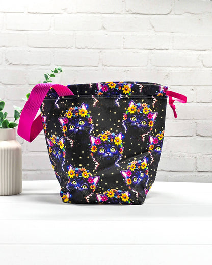 Projekttasche Tuva M aus schwarzem Baumwollstoff mit bunten Blumen-Katzen, pinkem Kordelzug und pinker Trageschlaufe. Handgenäht in Bayern von Nadelmikado. 