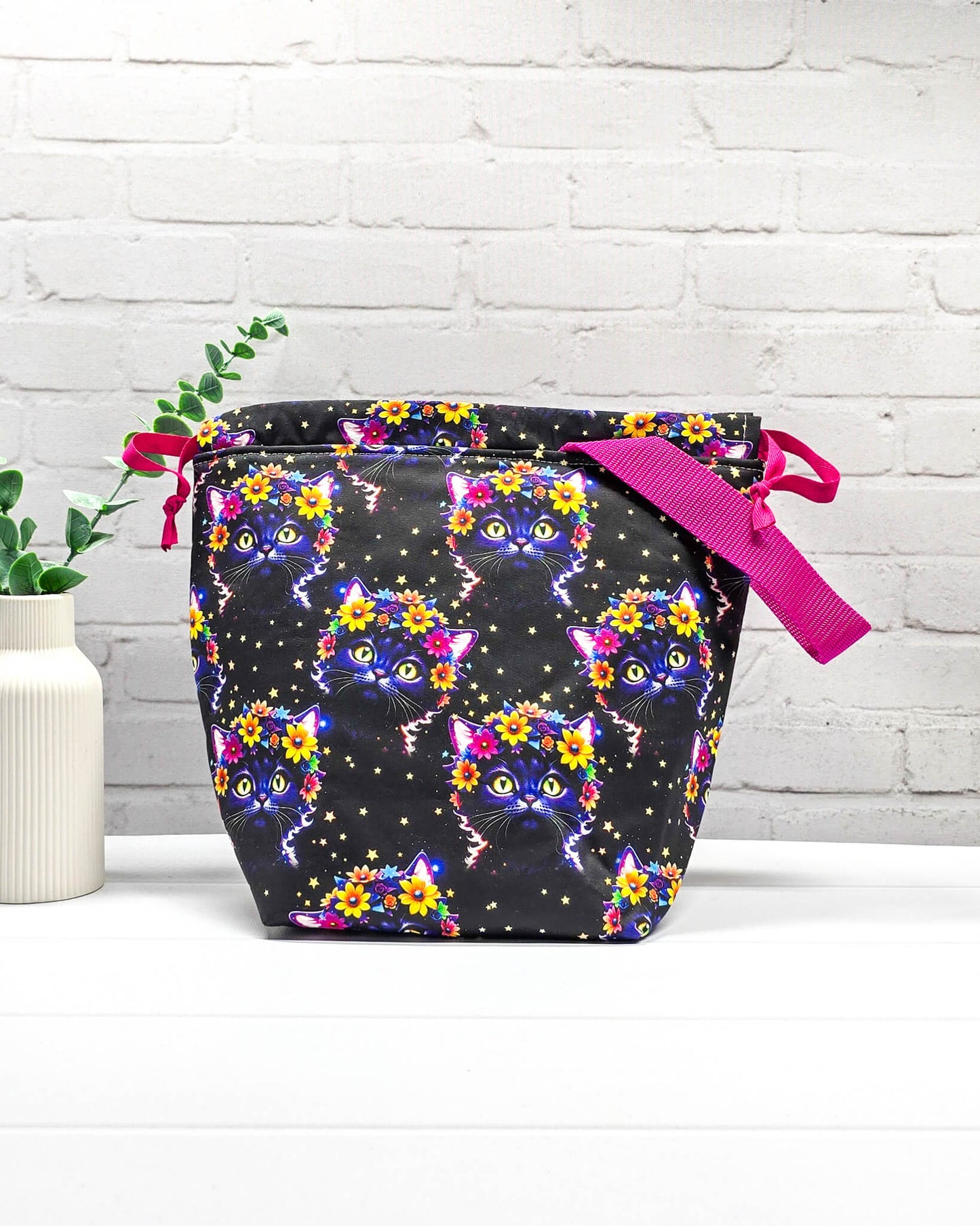 Projekttasche Tuva M aus schwarzem Baumwollstoff mit bunten Blumen-Katzen, pinkem Kordelzug und pinker Trageschlaufe. Handgenäht in Bayern von Nadelmikado. 