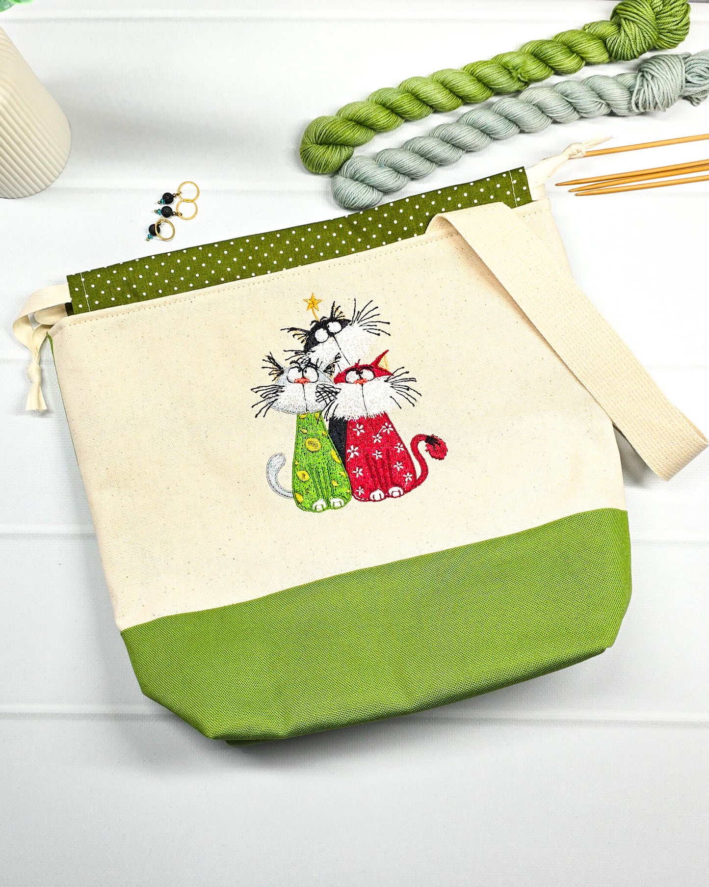 Projekttasche Tuva M aus hellem Canvas und grünem Stoff mit gesticktem Katzen-Trio (drei bunte Katzen mit Schnurrhaaren), grün weiß gepunktetem Kordelzug und cremefarbenem Tragegurt. Bietet Platz für 400g Wolle und schützt das Strickzeug vor Verheddern. Handgenäht in Bayern von Nadelmikado.